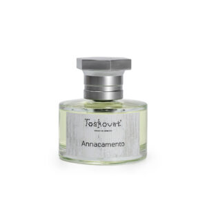 Perfume cilíndrico de líquido ámbar, tapa hexagonal gris, Annacamento en letras negras, etiqueta blanca.